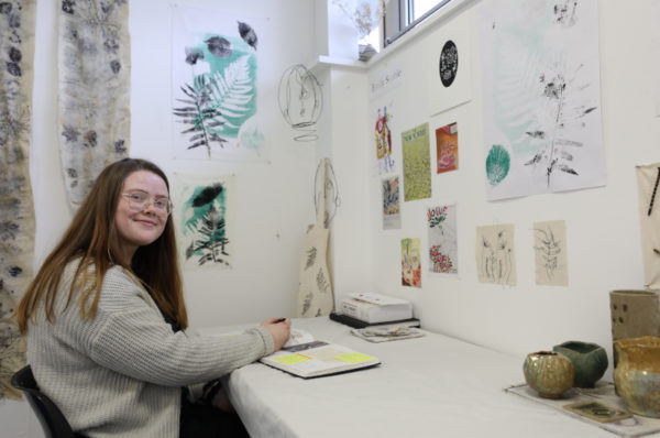 Student in her work space smiling at the camera while working on her sketchbook.