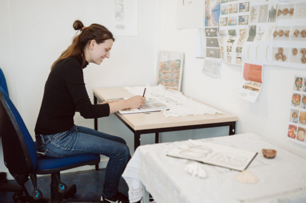 Smiling student drawing in her sketchbook sitting by a desk.