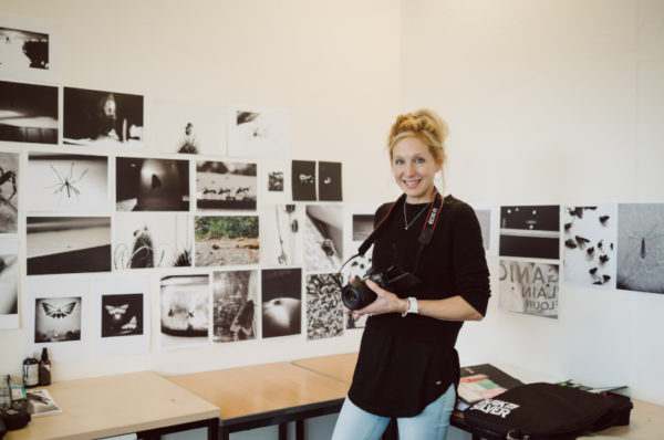 Student holding a camera in her work space and looking towards the person taking the picture.